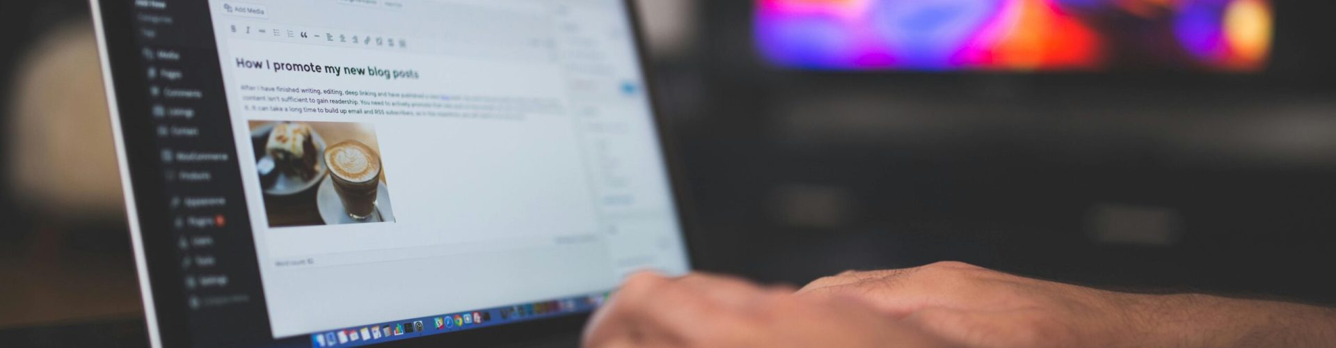 Hands typing on a laptop with a blog post visible, cozy indoor setting with colorful screen in background.